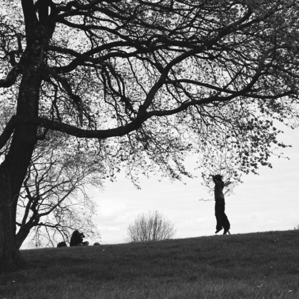 Photographie en noir et blanc d'un jardin en Écosse. On peut voir un grand arbre à gauche et sur la butte une jeune fille qui prend en photo une autre fille en train de sauter.