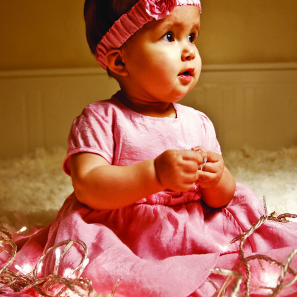 Portrait d'un bébé. Elle regarde par la fenêtre et la lumière vient éclairé son visage. Elle est assise sur une couverture blanche au long poile et habillé d'une jolie robe rose.