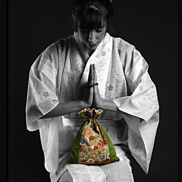 Photographie en noir et blanc d'une jeune femme en kimono qui joint les mains et se baisse pour saluer. Au centre sur les genoux de la Japonaise se trouve une bourse décorée de fleurs en couleur.